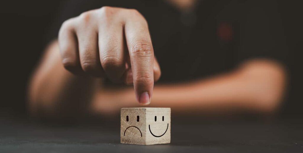 man hand points on a wooden cube with happy smile face on bright side and unhappy face on dark side of wooden block cube for a positive mindset selection. emotional state and mental health concept.