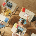 Top View Shot in Elementary School Computer Science Classroom: Children Sitting at their School Desk Using Personal Computers and Digital Tablets for Assignments.